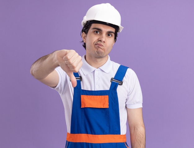 Hombre joven constructor en uniforme de construcción y casco de seguridad mirando al frente disgustado mostrando los pulgares hacia abajo de pie sobre la pared púrpura