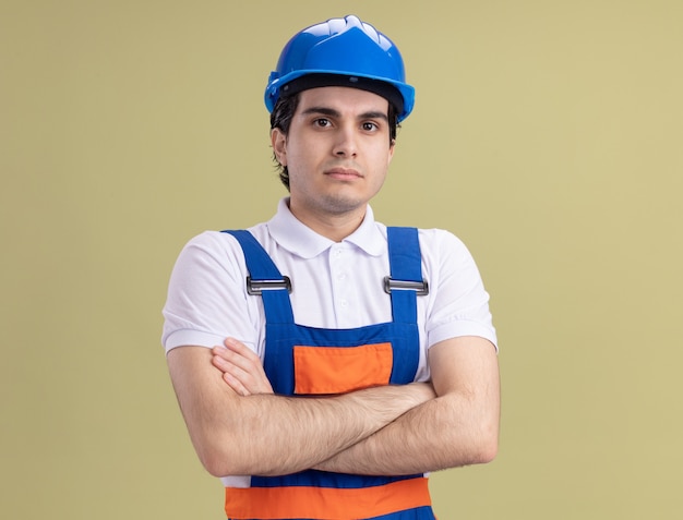 Hombre joven constructor en uniforme de construcción y casco de seguridad mirando al frente con expresión de confianza con los brazos cruzados de pie sobre la pared verde