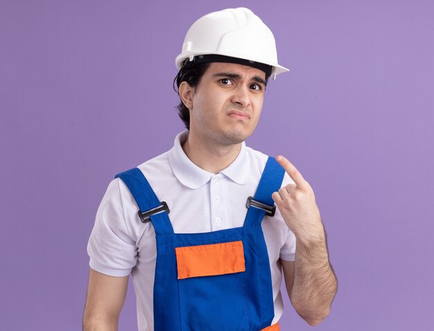 Hombre joven constructor en uniforme de construcción y casco de seguridad mirando al frente con expresión triste confundida apuntando a sí mismo de pie sobre la pared púrpura