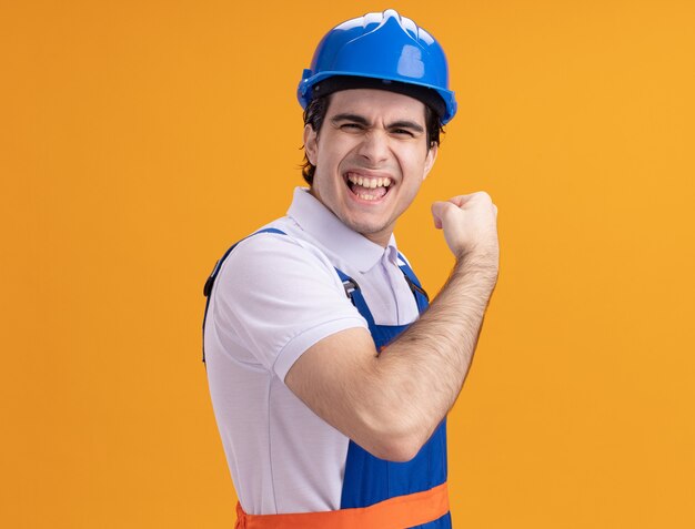 Hombre joven constructor en uniforme de construcción y casco de seguridad mirando al frente feliz y emocionado puño de pie sobre la pared naranja