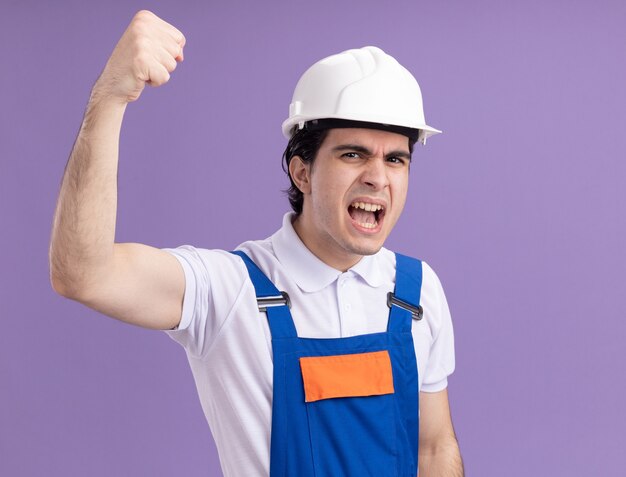 Hombre joven constructor en uniforme de construcción y casco de seguridad mirando al frente gritando con cara enojada levantando el puño de pie sobre la pared púrpura