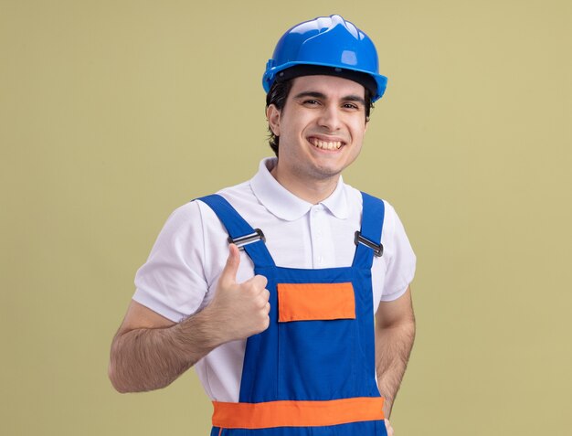 Hombre joven constructor en uniforme de construcción y casco de seguridad mirando al frente sonriendo alegremente mostrando los pulgares para arriba de pie sobre la pared verde