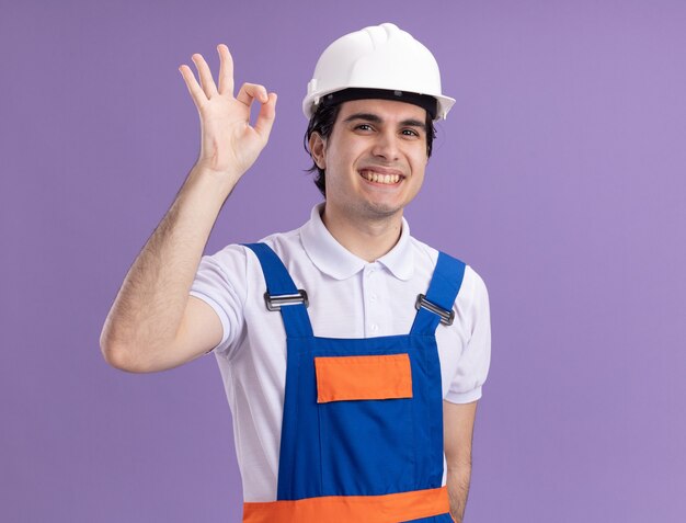 Hombre joven constructor en uniforme de construcción y casco de seguridad mirando al frente sonriendo alegremente mostrando signo ok de pie sobre la pared púrpura
