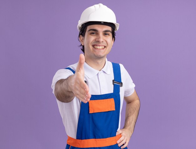 Hombre joven constructor en uniforme de construcción y casco de seguridad mirando al frente sonriendo amable ofreciendo saludo de mano de pie sobre la pared púrpura