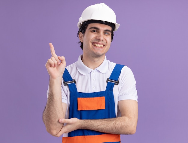 Hombre joven constructor en uniforme de construcción y casco de seguridad mirando al frente sonriendo apuntando con el dedo índice hacia el lado de pie sobre la pared púrpura
