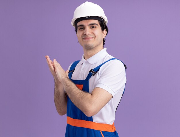 Hombre joven constructor en uniforme de construcción y casco de seguridad mirando al frente sonriendo confiado aplaudiendo de pie sobre la pared púrpura