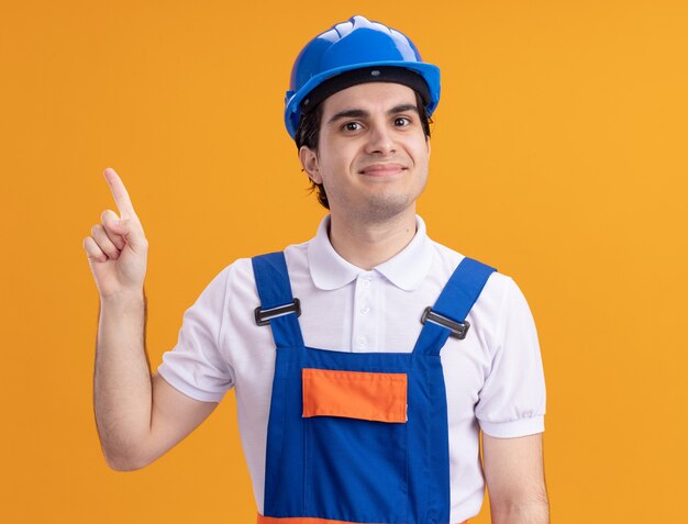 Hombre joven constructor en uniforme de construcción y casco de seguridad mirando al frente sonriendo confiado apuntando con el dedo índice de pie sobre la pared naranja