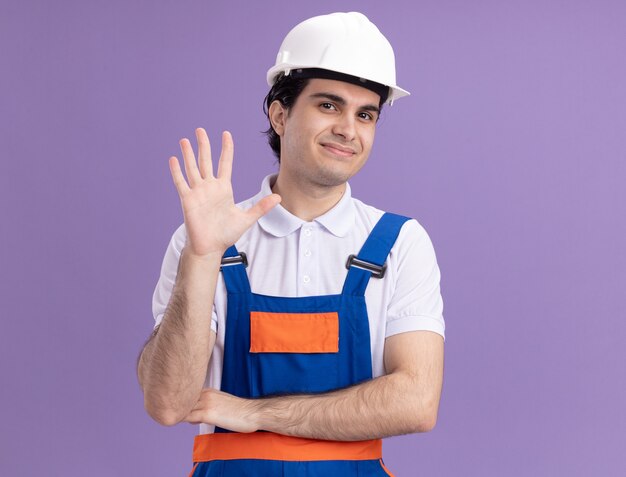 Hombre joven constructor en uniforme de construcción y casco de seguridad mirando al frente con una sonrisa en la cara saludando con la mano de pie sobre la pared púrpura