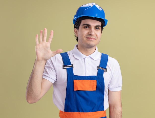 Hombre joven constructor en uniforme de construcción y casco de seguridad mirando al frente con una sonrisa en la cara saludando con la mano de pie sobre la pared verde