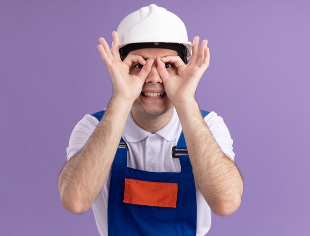 Hombre joven constructor en uniforme de construcción y casco de seguridad mirando al frente a través de los dedos haciendo gesto binocular sonriendo de pie sobre la pared púrpura