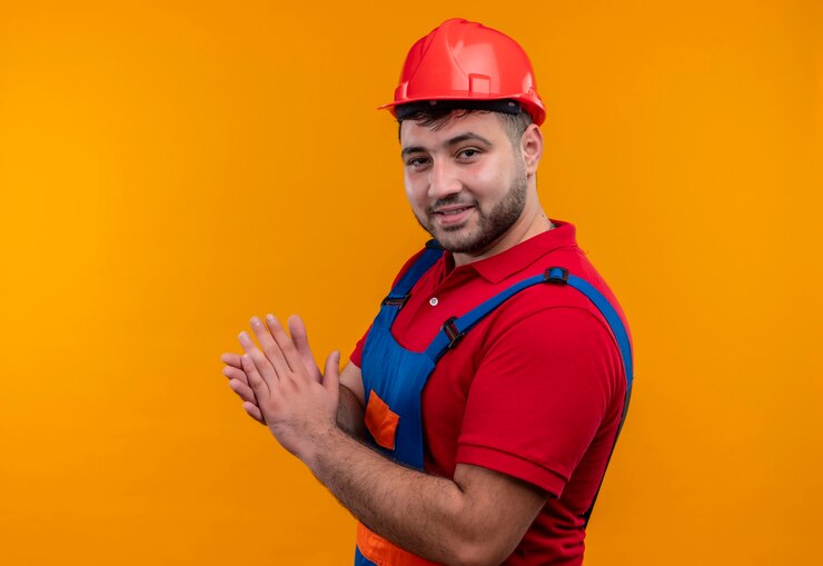 Foto gratuito hombre joven constructor en uniforme de construcción y casco de seguridad mirando a cámara aplaudiendo sonriendo confiado