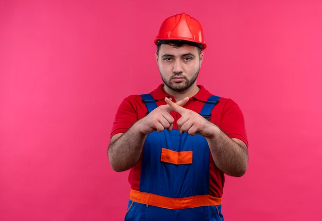 Hombre joven constructor en uniforme de construcción y casco de seguridad mirando a la cámara con el ceño fruncido haciendo gesto de defensa cruzando los dedos índices