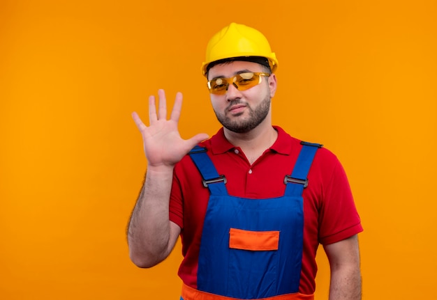 Hombre joven constructor en uniforme de construcción y casco de seguridad mirando a cámara saludando con la mano sonriendo