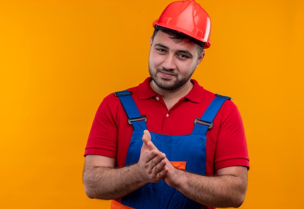 Foto gratuita hombre joven constructor en uniforme de construcción y casco de seguridad mirando a cámara con sonrisa de confianza frotando los brazos