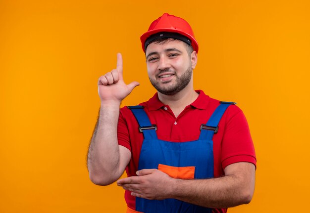 Hombre joven constructor en uniforme de construcción y casco de seguridad mirando confiado apuntando con el dedo índice hacia arriba