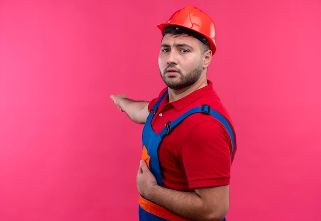 Foto gratuita hombre joven constructor en uniforme de construcción y casco de seguridad mirando confundido apuntando con el dedo índice a algo detrás