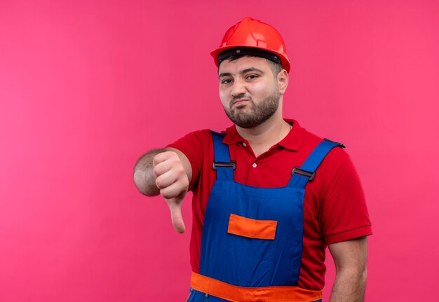 Hombre joven constructor en uniforme de construcción y casco de seguridad mirando disgustado mostrando los pulgares hacia abajo