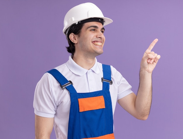 Hombre joven constructor en uniforme de construcción y casco de seguridad mirando a un lado sonriendo apuntando con el dedo índice a algo parado sobre la pared púrpura
