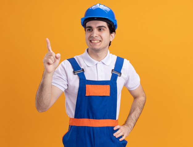 Hombre joven constructor en uniforme de construcción y casco de seguridad mirando a un lado sonriendo confiado apuntando con el dedo índice a algo parado sobre la pared naranja