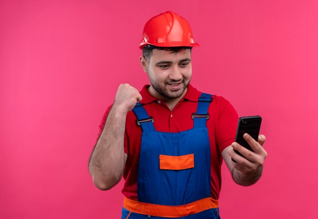 Hombre joven constructor en uniforme de construcción y casco de seguridad mirando la pantalla del teléfono inteligente apretando el puño feliz y salido regocijándose de su éxito