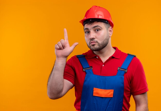 Hombre joven constructor en uniforme de construcción y casco de seguridad mirando sorprendido mostrando el dedo índice