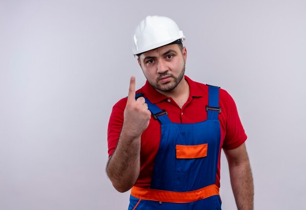Hombre joven constructor en uniforme de construcción y casco de seguridad mostrando el dedo índice con cara enojada
