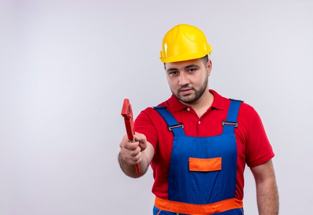 Hombre joven constructor en uniforme de construcción y casco de seguridad mostrando llave mirando a cámara disgustado