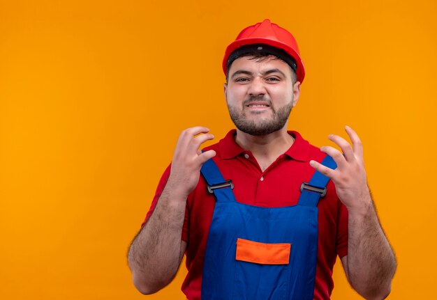 Hombre joven constructor en uniforme de construcción y casco de seguridad muy enojado, frustrado y estresado levantando los brazos