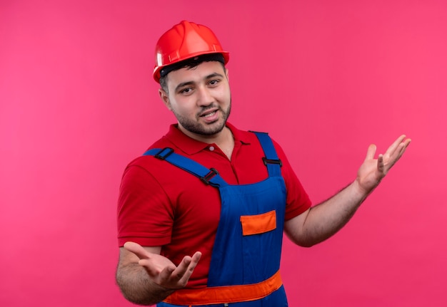 Hombre joven constructor en uniforme de construcción y casco de seguridad que parece confundido e incierto encogiéndose de hombros sin respuesta