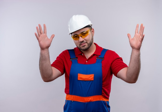 Hombre joven constructor en uniforme de construcción y casco de seguridad que parece confundido e incierto encogiéndose de hombros sin respuesta