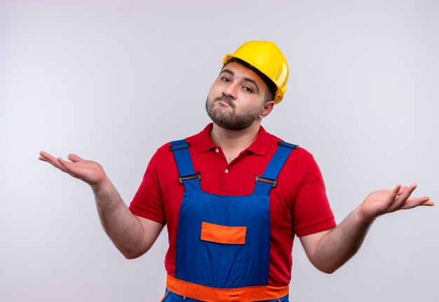 Hombre joven constructor en uniforme de construcción y casco de seguridad que parece confundido e incierto encogiéndose de hombros sin respuesta