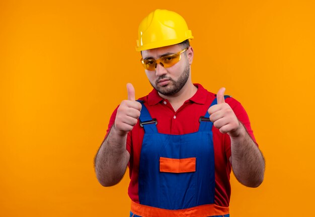 Hombre joven constructor en uniforme de construcción y casco de seguridad que parece seguro mostrando los pulgares hacia arriba con ambas manos