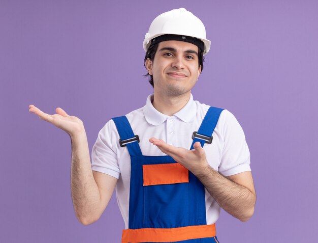 Hombre joven constructor en uniforme de construcción y casco de seguridad que presenta espacio de copia con los brazos sonriendo alegremente de pie sobre la pared púrpura