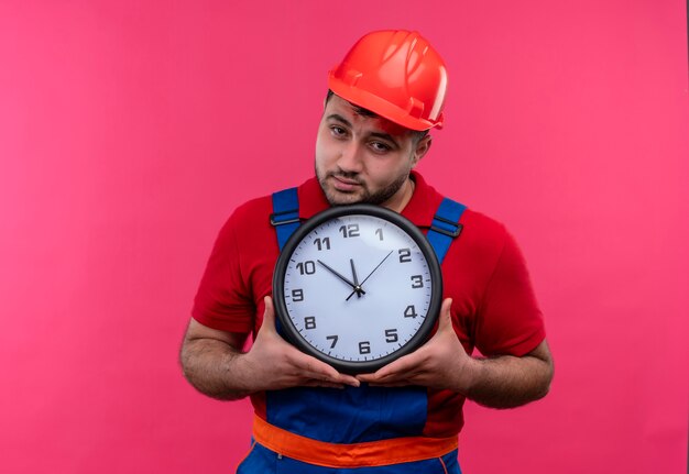 Hombre joven constructor en uniforme de construcción y casco de seguridad con reloj de pared mirando a cámara con cara infeliz