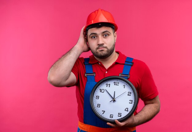 Hombre joven constructor en uniforme de construcción y casco de seguridad con reloj de pared mirando confundido y sorprendido