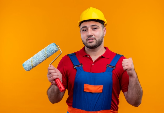 Hombre joven constructor en uniforme de construcción y casco de seguridad con rodillo de pintura apretando el puño como un ganador