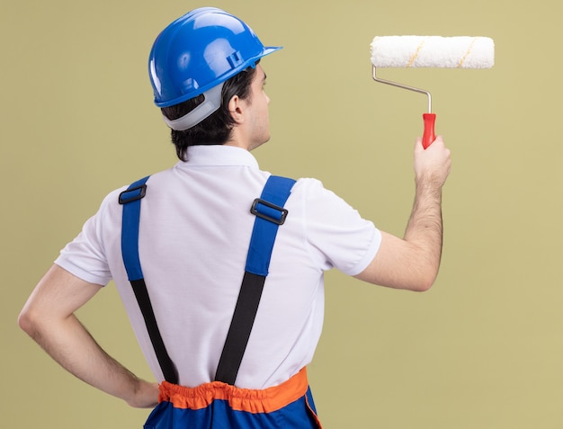 Foto gratuita hombre joven constructor en uniforme de construcción y casco de seguridad con rodillo de pintura de pie con la espalda sobre la pared verde