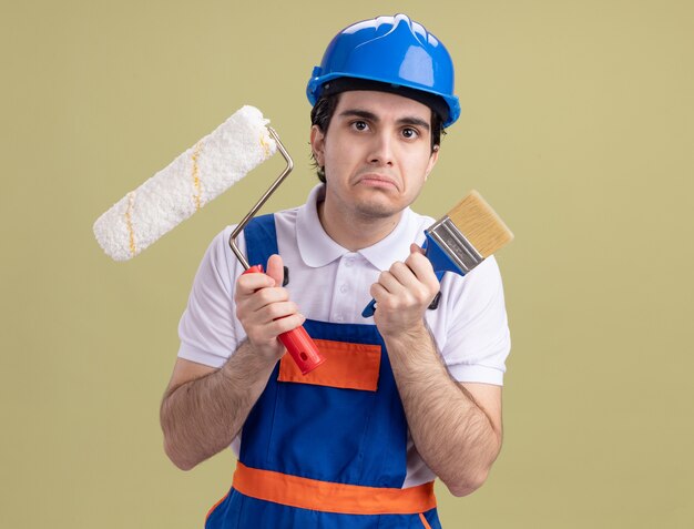 Hombre joven constructor en uniforme de construcción y casco de seguridad con rodillo de pintura y pincel mirando al frente con expresión triste frunciendo los labios de pie sobre la pared verde