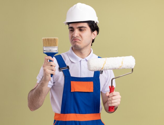 Hombre joven constructor en uniforme de construcción y casco de seguridad con rodillo de pintura y pincel que parece confundido teniendo dudas sobre la pared verde