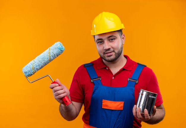 Hombre joven constructor en uniforme de construcción y casco de seguridad con rodillo de pintura y pintura puede sonriendo