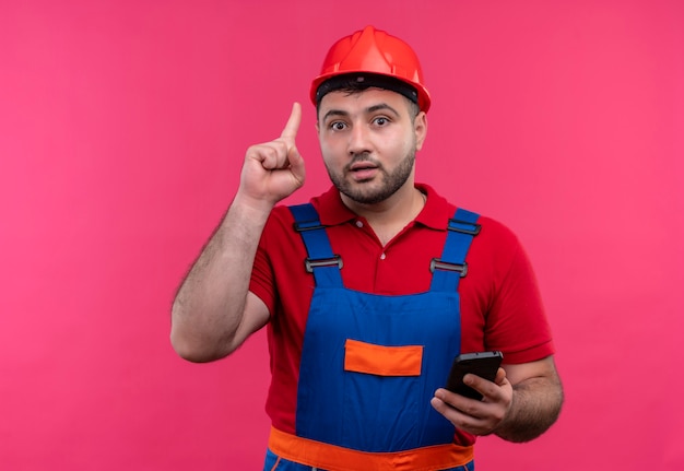 Hombre joven constructor en uniforme de construcción y casco de seguridad con smartphone mostrando el dedo índice recordándose a sí mismo que no debe olvidar