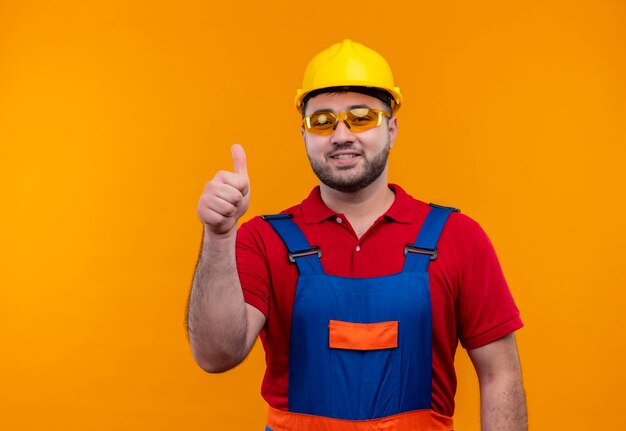 Hombre joven constructor en uniforme de construcción y casco de seguridad sonriendo alegremente mostrando los pulgares para arriba