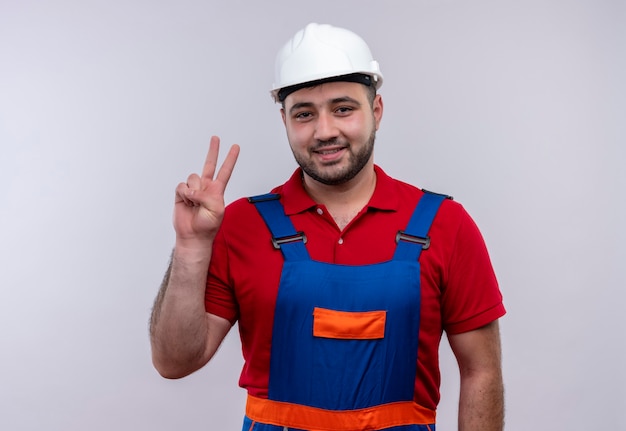 Hombre joven constructor en uniforme de construcción y casco de seguridad sonriendo mostrando y apuntando hacia arriba con los dedos número dos