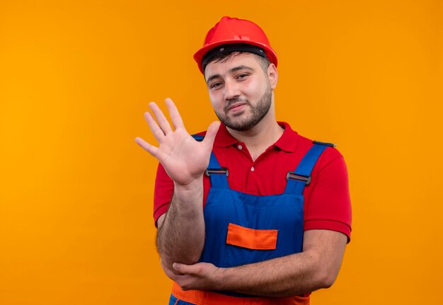 Hombre joven constructor en uniforme de construcción y casco de seguridad sonriendo saludando con la mano