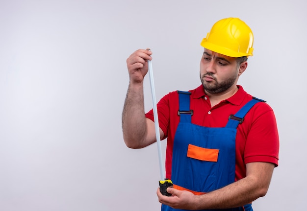 Hombre joven constructor en uniforme de construcción y casco de seguridad sosteniendo cinta métrica mirándolo con cara seria