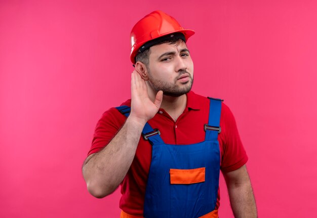 Hombre joven constructor en uniforme de construcción y casco de seguridad sosteniendo la mano cerca de la oreja tratando de escuchar