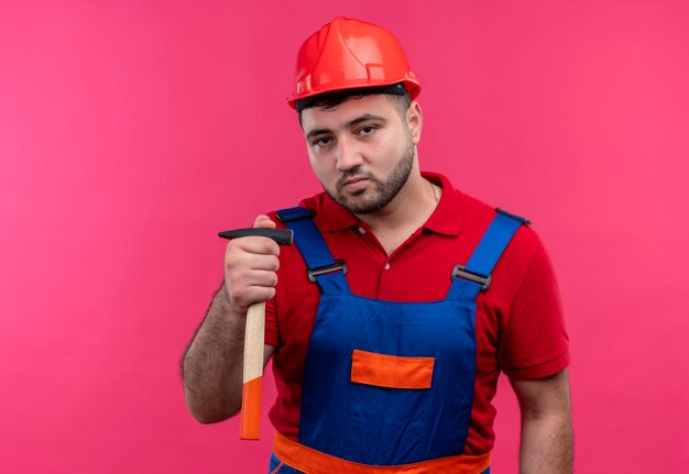 Hombre joven constructor en uniforme de construcción y casco de seguridad sosteniendo un martillo en la mano mirando confiado