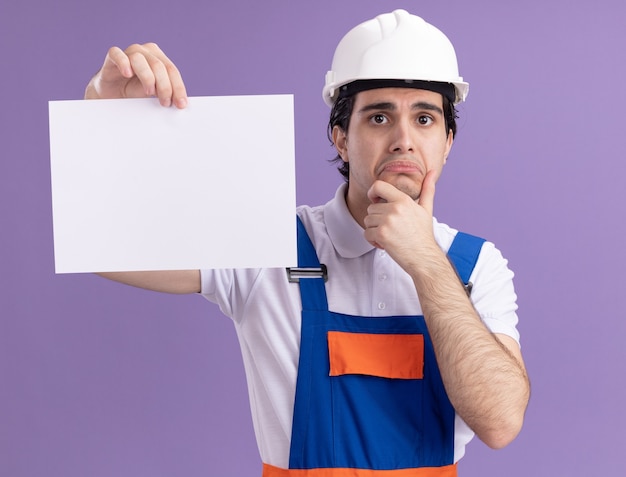 Hombre joven constructor en uniforme de construcción y casco de seguridad sosteniendo una página en blanco mirando al frente con expresión triste frunciendo los labios de pie sobre la pared púrpura