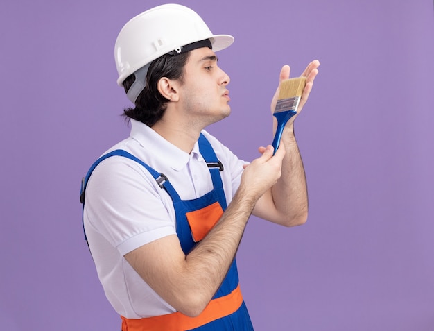 Hombre joven constructor en uniforme de construcción y casco de seguridad sosteniendo pincel mirándolo con amor de pie sobre la pared púrpura