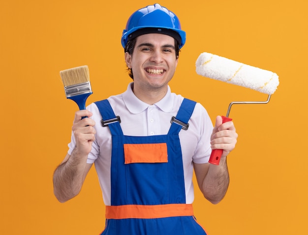 Foto gratuita hombre joven constructor en uniforme de construcción y casco de seguridad sosteniendo pincel y rodillo mirando al frente sonriendo alegremente de pie sobre la pared naranja